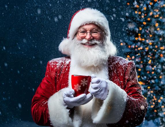 Santa standing in front of a Christmas tree and holding a festive mug. The background is snowy.