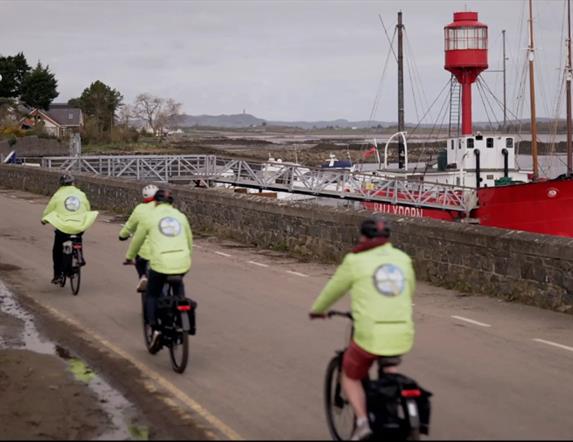 A group of cyclists in hi-vis jackets cycle along a coastal road. A red boat is docked at the shore.