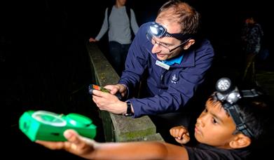 Child wearing a head torch and holding out a bat detector, guided by a member of staff. 