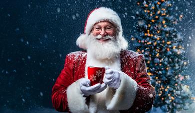 Santa standing in front of a Christmas tree and holding a festive mug. The background is snowy.