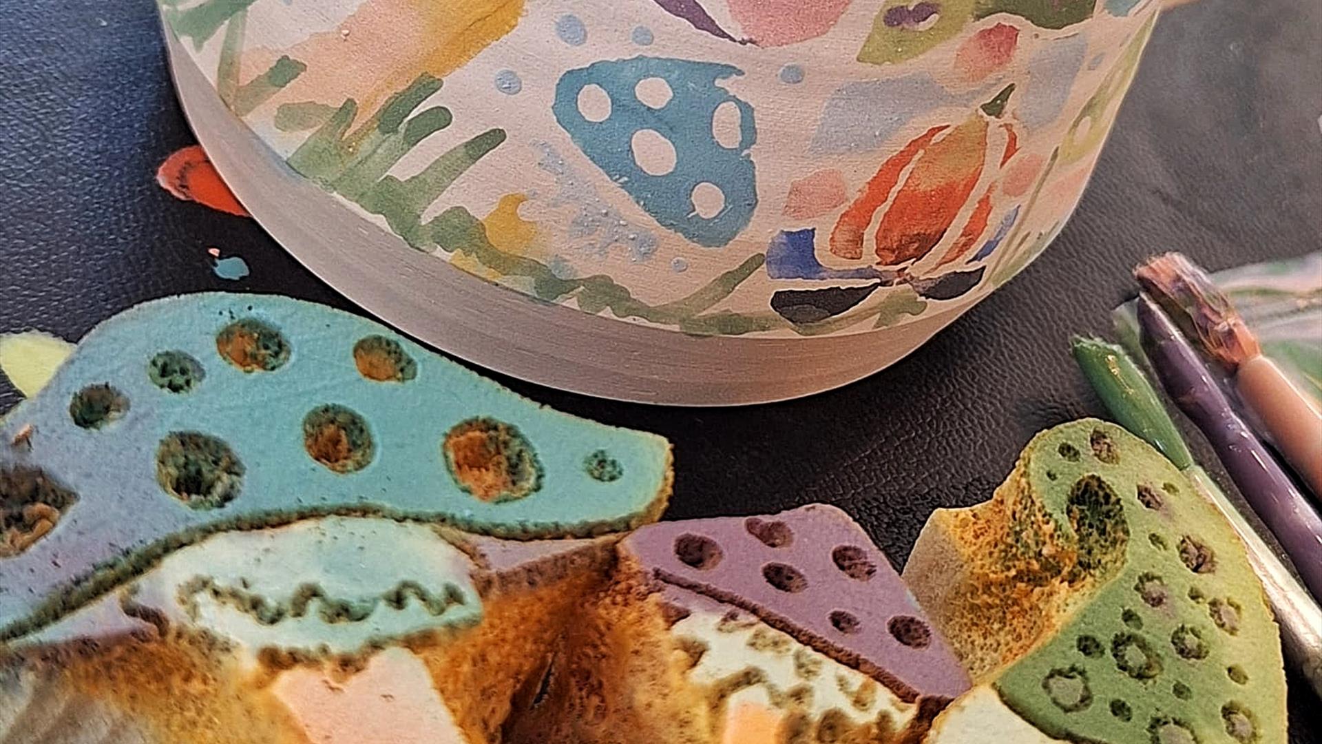 Hand thrown pottery mug in the background, decorated in colourful sponge prints of gnomes and toadstools. The sponge cut outs are in front of the mug,