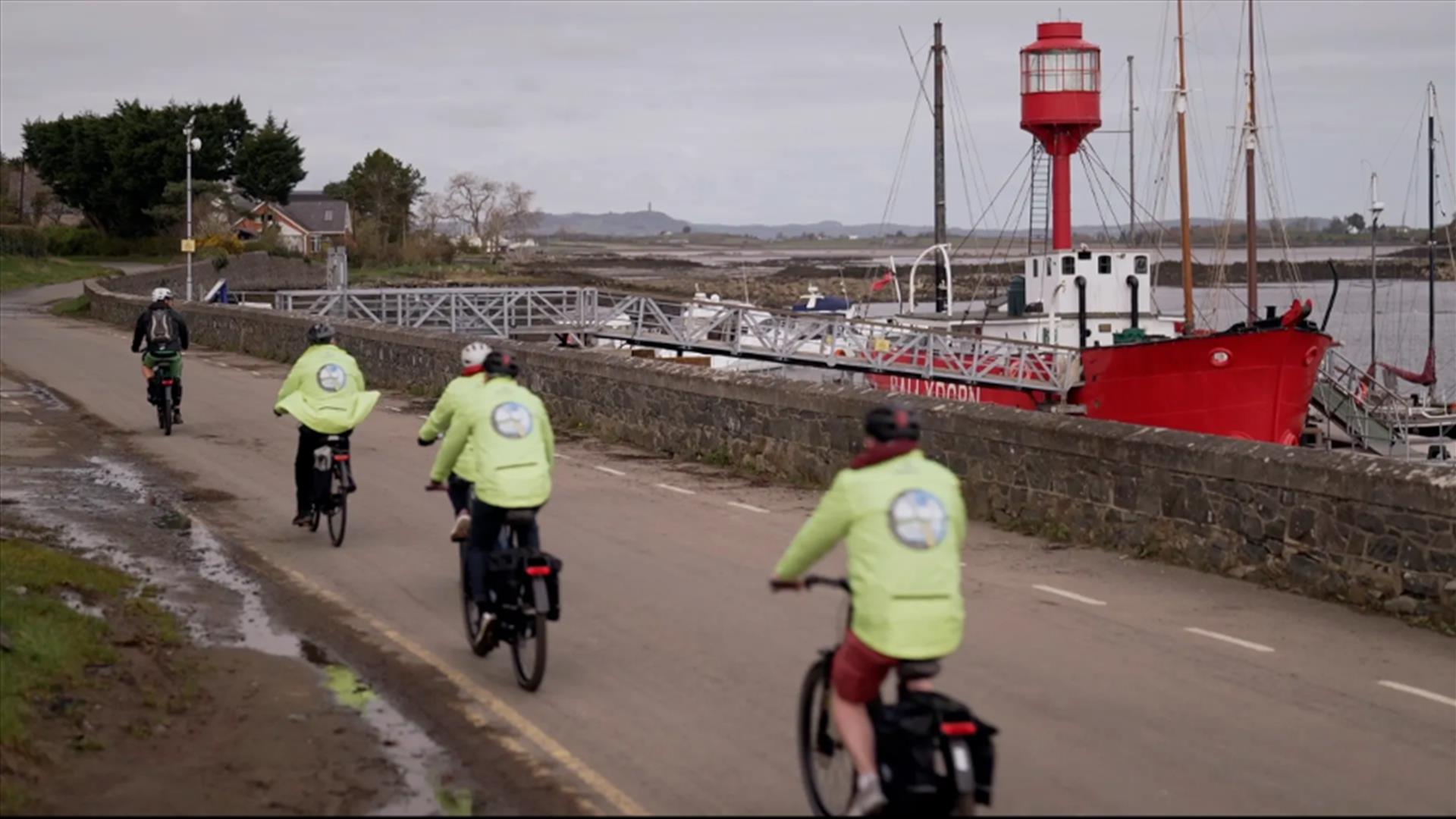 A group of cyclists in hi-vis jackets cycle along a coastal road. A red boat is docked at the shore.