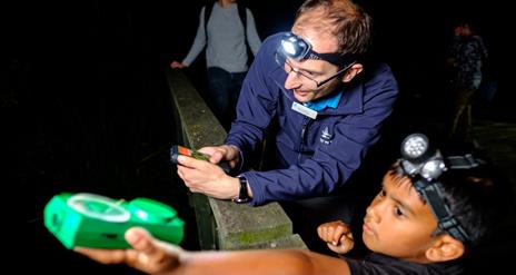 Child wearing a head torch and holding out a bat detector, guided by a member of staff.