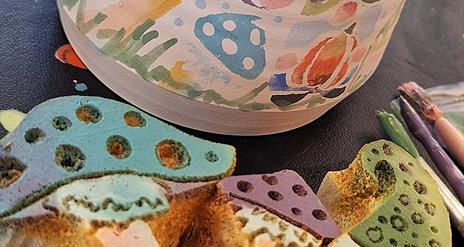 Hand thrown pottery mug in the background, decorated in colourful sponge prints of gnomes and toadstools. The sponge cut outs are in front of the mug,