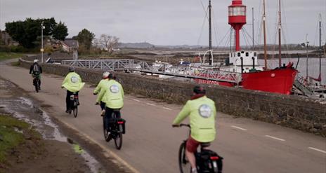 A group of cyclists in hi-vis jackets cycle along a coastal road. A red boat is docked at the shore.